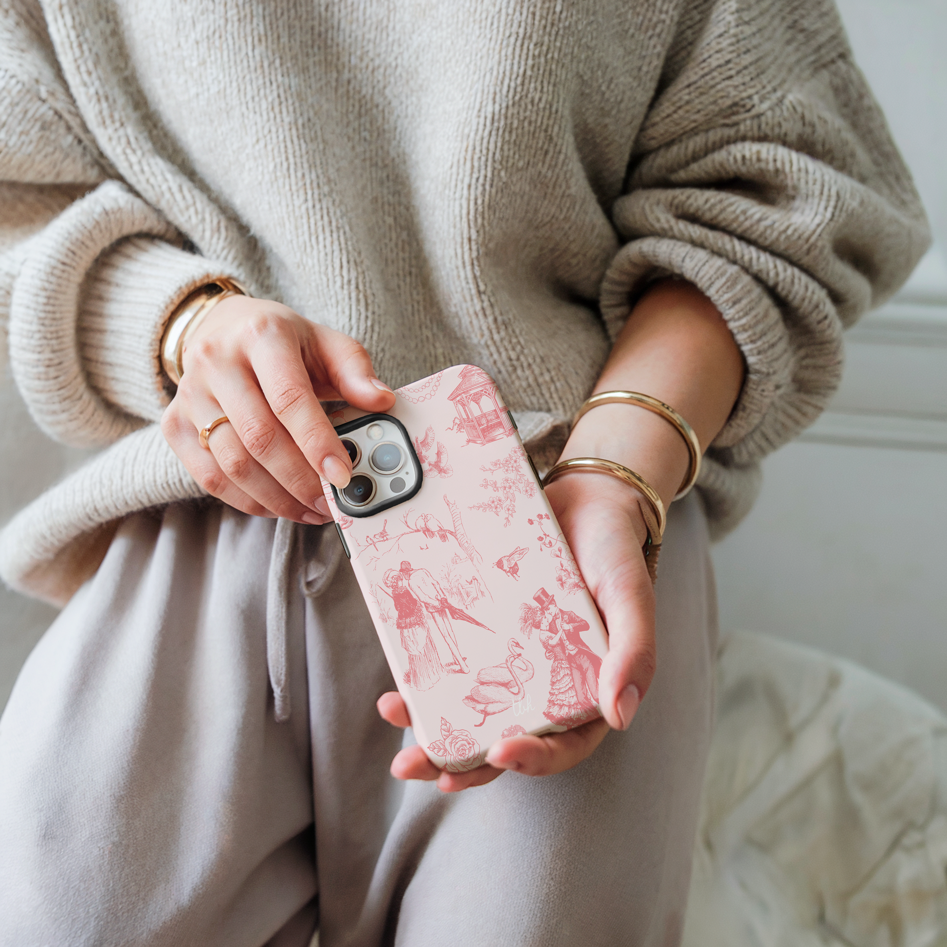 Person holding a phone with a pink case featuring vintage designs, wearing a beige sweater and pants.